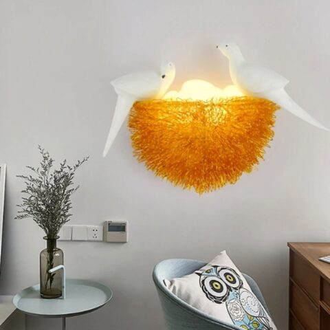 En el centro de la imagen hay una aplicación mural con forma de nido de pájaro. En la parte inferior izquierda de la imagen hay una mesa con un jarrón gris transparente con ramas en su interior. En la esquina inferior derecha se ve la parte superior de un sillón con un cojín de búho.