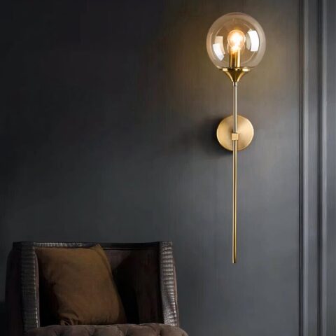 Un aplique moderno y elegante compuesto por un esbelto tallo dorado sobre el que hay una bola de cristal con una bombilla en su interior. El tallo se fija a la pared mediante una base redonda dorada. Sobre una pared negra. A su lado hay un sillón, del que sólo se ve el respaldo marrón.