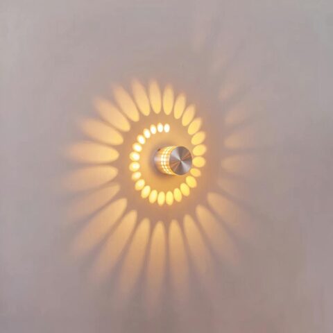 Aplique LED circular de aluminio plateado encendido, proyecta una espiral de luz cálida sobre una pared blanca