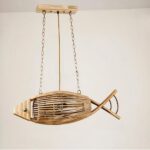 Araña de madera en forma de pez, suspendida por dos cadenas en cada extremo. Conectada a una base rectangular de madera. Detrás hay una pared beige.