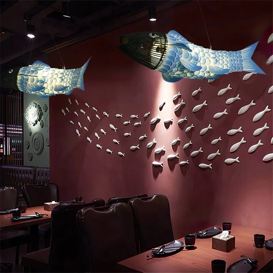 Sala de restaurante con dos lámparas de araña en forma de grandes peces con cuerpos azules y blancos y cabezas hechas con palos de madera negra. Hay mesas dispuestas y una pared roja al fondo con muchos peces colgados de la pared como un banco de peces en el mar.