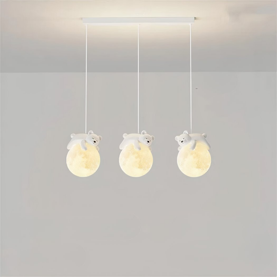 Lámpara colgante formada por tres cables, cada uno con una bola que representa la luna y un osito de peluche durmiendo sobre la luna. Las bolas están iluminadas. La base rectangular cuelga del techo gris claro, con una pared gris claro de fondo.