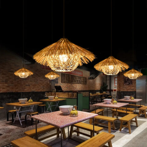 Una lámpara de araña de paja natural al estilo de una exótica cabaña de paja, colgada de la pared e iluminada, encima de las mesas de lo que parece ser un restaurante, en un ambiente de iluminación tenue.