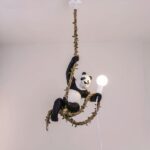 Suspensión con cuerda de cáñamo entrelazada con follaje. Un panda cuelga de su pata delantera derecha, la cuerda pasa por sus patas traseras y sujeta una bombilla en su pata delantera izquierda con parte de la cuerda.